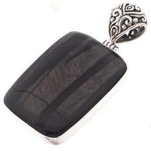 Peut inclure: Un pendentif rectangulaire avec une pierre polie gris foncé présentant des motifs linéaires. La pierre est sertie dans un cadre en métal argenté et possède une bélière ornée d'un motif tourbillonnant. Un bijou.