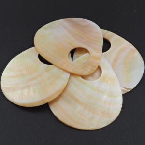 May include: Four pearlescent, teardrop-shaped shell pendants with a hole near the top. The shells have a natural, swirling pattern of cream, peach, and pale yellow hues. The pendants are arranged on a dark background.