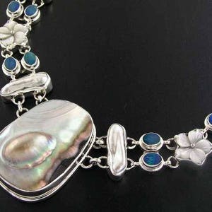 以下が含まれることがあります： 虹色のアワビの貝殻の大きなペンダント、白い花のアクセント、そして楕円形の青いオパールストーンが付いたシルバーネックレスです。