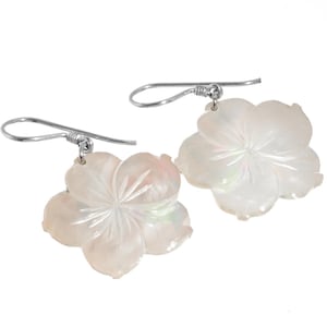 May include: A pair of flower-shaped earrings. Each earring features a carved, white, mother-of-pearl flower pendant with five petals. The earrings have silver-colored, curved fish hook ear wires. The earrings are designed to be worn by anyone.