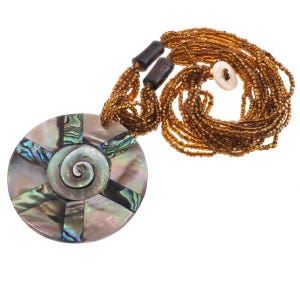 May include: A necklace featuring a large circular pendant made of iridescent abalone shell segments arranged in a radial pattern with a central spiral. The necklace has multiple strands of small, translucent amber-colored beads and a white button clasp.