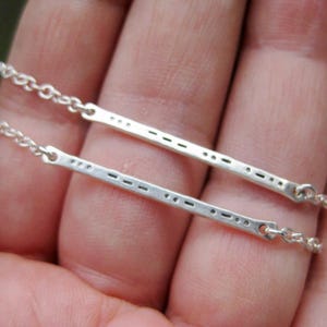 May include: Two silver-toned metal bar necklaces featuring a series of small dots and dashes engraved along the surface. Each thin rectangular bar is attached to a delicate silver-toned chain on both ends, creating a horizontal pendant design.