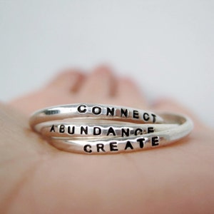 The photo shows a silver ring resting on an open hand. The ring has 3 separate rings linked together and each ring has a word stamped on it. Connect, abundance and create are the three words.
