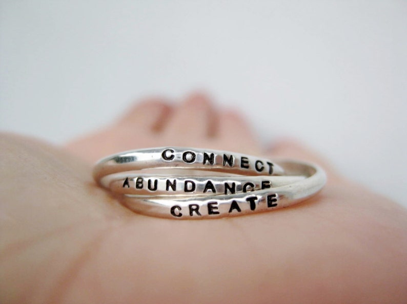 The photo shows a silver ring resting on an open hand. The ring has 3 separate rings linked together and each ring has a word stamped on it. Connect, abundance and create are the three words.