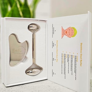May include: A stainless steel facial roller and gua sha tool set, presented in a white box. The set includes a roller with two rounded ends and a heart-shaped gua sha tool. The box also features instructions and the text "Bella Face".