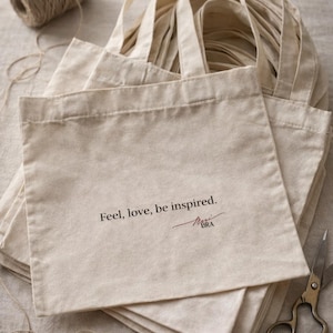 Puede incluir: Un mont&oacute;n de bolsas de mano de algod&oacute;n beige con asas, cada una impresa con las palabras "Feel, love, be inspired." y una firma. Tambi&eacute;n se ven un carrete de hilo y unas tijeras. Las bolsas forman parte de una l&iacute;nea de embalaje sostenible.