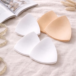 Op de afbeelding: Vier driehoekige bh-inzetstukken in de afbeelding. Twee zijn wit en twee zijn lichtbeige van kleur. De inzetstukken zijn gerangschikt op een neutraal gekleurde stof, met een meetlint en een gedeeltelijk zichtbaar open boek op de achtergrond.