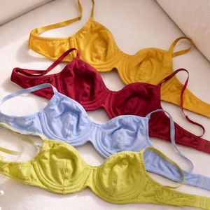 May include: A collection of four underwire bras in mustard yellow, deep burgundy, light periwinkle blue, and lime green. Each bra features adjustable shoulder straps, a structured underwire cup design, and a hook-and-eye closure on the back band.