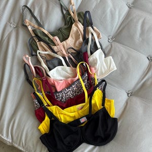 May include: A collection of multiple underwire bras stacked vertically, featuring a variety of colours including black, bright yellow, deep red, leopard print, soft pink, white, charcoal grey, beige, and olive green, all with adjustable shoulder straps.
