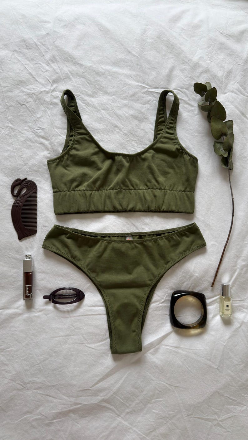 May include: Olive green lingerie set with a matching bralette and high-cut briefs. Accessories include a brown comb, lipstick, hair clip, bracelet, and perfume bottle. A sprig of eucalyptus adds a natural touch.
