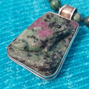 May include: A rectangular pendant featuring a carved frog made of green zoisite with black inclusions and a prominent magenta ruby patch. The stone is set in a silver frame with a silver bail, attached to a beaded necklace of round green stones.