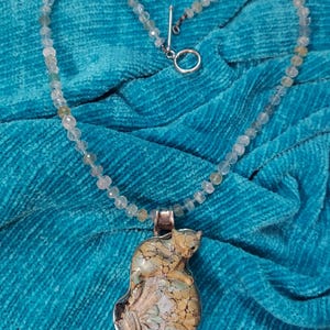 Può includere: Una collana con una fila di piccole perline sfaccettate e traslucide nei toni del verde pallido e trasparente, rifinita con una chiusura a T argentata. Il ciondolo è un pezzo sagomato con un motivo screpolato e marmorizzato beige, verde e crema.
