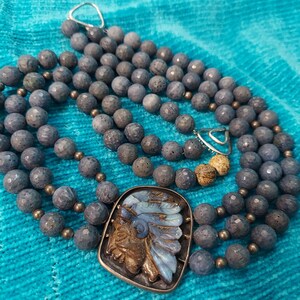 May include: A multi-strand necklace featuring gray-blue stone beads and a silver pendant. The pendant has a detailed design of a Native American chief with blue and brown accents. The necklace is displayed on a turquoise fabric.