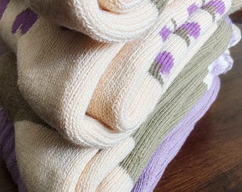 May include: A stack of three pairs of socks in different colors. The top pair is beige with purple flowers, the middle pair is beige with green stripes, and the bottom pair is purple.