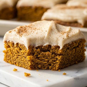 May include: A square piece of golden-brown pumpkin spice cake topped with a thick layer of light beige cream cheese frosting and a light dusting of cinnamon. The cake has a porous, moist texture and a visible bite mark on the front edge.