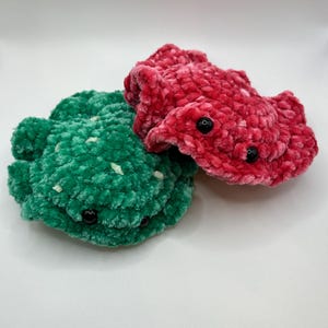May include: Two handmade crochet creatures in the image. One is green with white flecks, the other is red. Both have black button eyes. The plush toys are made of a soft, textured yarn.