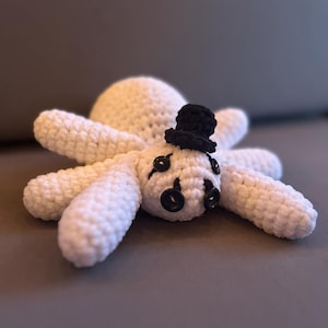 May include: A white crocheted spider plush toy featuring eight legs, a round body, and a small black top hat. The face is decorated with four black button eyes arranged in a cluster, providing a distinct look to this handmade stuffed creature.