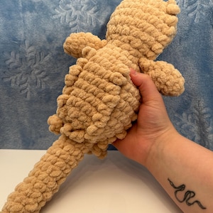 Può includere: Bambola all'uncinetto beige fatta a mano con un aspetto strutturato e morbido. La bambola ha una testa arrotondata, braccia piccole e una lunga coda cilindrica. La bambola è tenuta su uno sfondo blu.