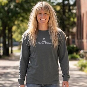 May include: A charcoal gray long-sleeve t-shirt featuring the white text I throw things on the chest, paired with blue denim jeans and white sneakers. The shirt has a crew neckline and long sleeves, while the jeans have a classic straight-leg fit.