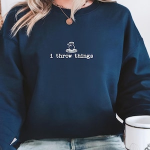 May include: A navy blue crewneck sweatshirt featuring a white line art graphic of a pottery wheel with a vase and the text "i throw things" printed in a typewriter-style font centered on the chest area of the garment.