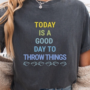 May include: A charcoal gray short-sleeve t-shirt featuring the text "TODAY IS A GOOD DAY TO THROW THINGS" in yellow, green, and blue, with a row of six light blue coffee cup icons printed along the bottom of the graphic design.