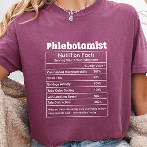 May include: A maroon t-shirt featuring the word "Phlebotomist" and "Nutrition Facts" in white text. The graphic lists daily values for phlebotomy skills, including tourniquet skills, small talk, and pain distraction.