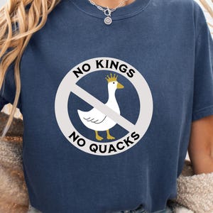 May include: Navy blue t-shirt with a white graphic of a goose wearing a crown, crossed out by a red line. The text reads "NO KINGS NO QUACKS" in a circular design around the goose.