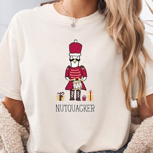 May include: Cream-colored t-shirt featuring a cartoon duck dressed as a nutcracker soldier, complete with a red uniform and a drum. The text "NUTQUACKER" is printed below the duck, with small gift boxes on either side.