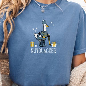 May include: A blue t-shirt featuring a cartoon goose dressed as a nutcracker soldier, complete with a hat, boots, and a staff. The text "NUTQUACKER" is printed below the goose, with snowflake and gift box graphics.
