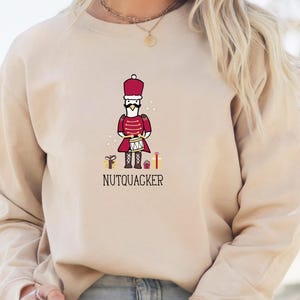 May include: Beige sweatshirt featuring a cartoon nutcracker goose character in a red uniform, holding a drum. The word "NUTQUACKER" is printed below the character, with gift box and star graphics.
