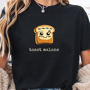 May include: Black t-shirt featuring a cartoon illustration of a slice of toast with a smiling face. The text "toast malone" is printed below the toast image in white, sans-serif font.
