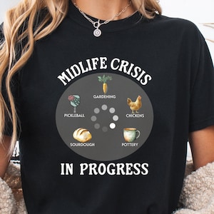 May include: A black t-shirt featuring a graphic with the text MIDLIFE CRISIS IN PROGRESS. The design includes a loading icon surrounded by illustrations of a carrot, a chicken, a mug, a loaf of bread, and a pickleball paddle with labels.