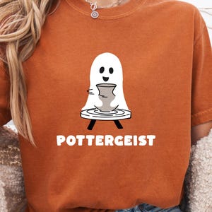 Peut inclure: Un t-shirt orange brûlé avec un motif de fantôme blanc travaillant sur un tour de potier. Le fantôme façonne un petit récipient en argile grise. Sous le motif, le texte POTTERGEIST est imprimé en lettres capitales blanches et grasses.