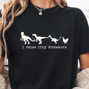 Puede incluir: Camiseta negra con un gráfico blanco de dinosaurios y una gallina, con el texto "i raise tiny dinosaurs". El diseño está impreso en una camiseta de cuello redondo.