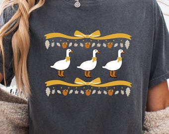 May include: Dark gray t-shirt featuring a whimsical design of three white geese with yellow bow ties, set against a backdrop of yellow ribbons, acorns, and leaves. The design is centered on the shirt, creating a festive and autumnal theme.