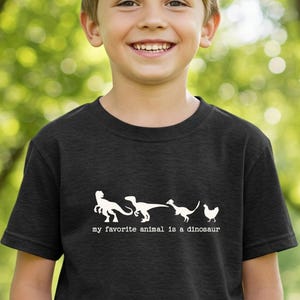 Puede incluir: Camiseta negra con un gráfico blanco de siluetas de dinosaurios y una gallina. El texto "my favorite animal is a dinosaur" está debajo. La camiseta la lleva un niño sonriente.