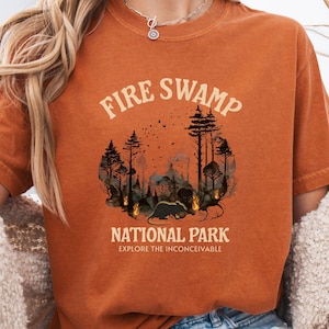 May include: Burnt orange t-shirt with a graphic design. The design features the text "FIRE SWAMP NATIONAL PARK" above a forest scene with flames and a creature. The phrase "EXPLORE THE INCONCEIVABLE" is below the park name.