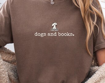 Può includere: Una maglietta marrone con la scritta "dogs and books". Sopra il testo c'è una piccola grafica di un cane seduto su un libro. La maglietta è tinta unita e ha uno scollo tondo.