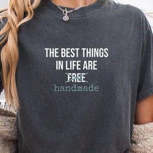 May include: Dark gray t-shirt with the text "THE BEST THINGS IN LIFE ARE handmade." The word "FREE" is crossed out. The shirt is a crew neck and has a vintage, worn look.