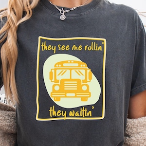 Puede incluir: Camiseta gris oscuro con un dibujo de autobús escolar amarillo dentro de un rectángulo con borde amarillo. El texto "they see me rollin' they waitin'" está impreso encima y debajo de la ilustración del autobús. Prenda de vestir informal.
