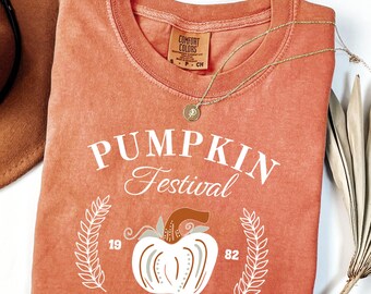 Op de afbeelding: Een koraalkleurig t-shirt met de woorden "PUMPKIN Festival" in witte letters en een pompoenafbeelding. Het shirt heeft ook de tekst "1982 LOCALLY GROWN". Een gouden ketting en een bruine hoed zijn ook zichtbaar.