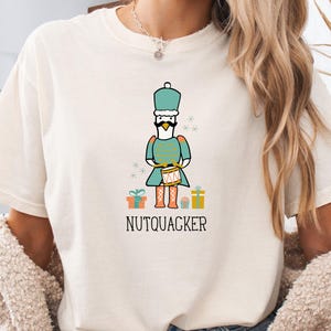 May include: Cream-colored t-shirt featuring a whimsical illustration of a nutcracker duck character in a teal uniform, holding a drum. The word "NUTQUACKER" is printed below the character, with gift boxes on either side. A fun, festive design.