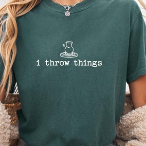 Op de afbeelding: Een donker bosgroen t-shirt met korte mouwen voorzien van een witte lijntekening van een pottenbakkersschijf met een vaas en de tekst "i throw things" gedrukt in een typemachine-stijl lettertype in het midden op de borst.