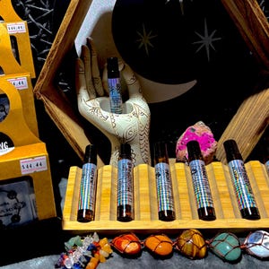 May include: A display of essential oil roller bottles with colorful labels, arranged on a wooden rack. The bottles are brown with silver and blue labels. A decorative hand holds a bottle. Gemstones and crystals are in the foreground.