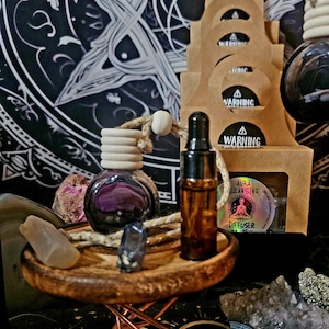 May include: A small round glass diffuser bottle with a wooden cap and hanging cord, a brown glass dropper bottle, and two small crystals on a wooden stand with a copper-colored wire base. Behind are stacked cardboard boxes with warning labels.