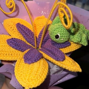 May include: A handmade crochet flower bouquet featuring a large yellow and purple flower with a green caterpillar. The bouquet is wrapped in purple tissue paper and tied with a lavender ribbon. The crochet work is detailed.