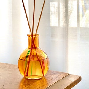 May include: A clear glass diffuser bottle with a round, textured amber base. The bottle is filled with a light yellow liquid and has four wooden sticks inserted into it.