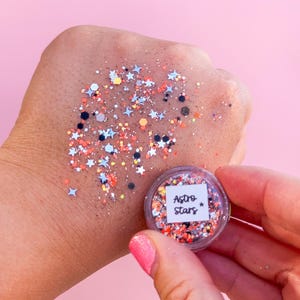 May include: A small round clear container of loose cosmetic glitter labeled Astro Stars, featuring a mix of orange, black, and silver hexagonal particles along with small silver star-shaped sequins, with a sample of the glitter spread out.