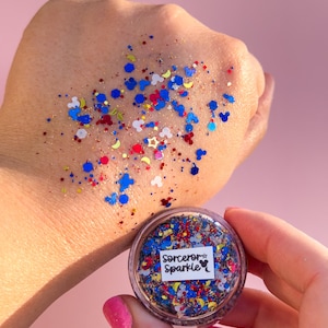 May include: A small, clear plastic pot filled with colourful glitter, including blue, red, white, and gold. The glitter is scattered on a hand, with the pot held in the other hand. The pot has a label that reads "Sorcerer Sparkle."