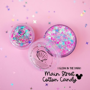Puede incluir: Tres frascos de plástico transparente llenos de purpurina colorida, incluyendo tonos rosas, azules y morados, están dispuestos sobre un fondo rosa. Los frascos tienen etiquetas con un diseño de concha y el texto "Facial Glitter" y "Hair Glitter". También es visible el texto "I Glow in the Dark!" y "Main Street Cotton Candy".